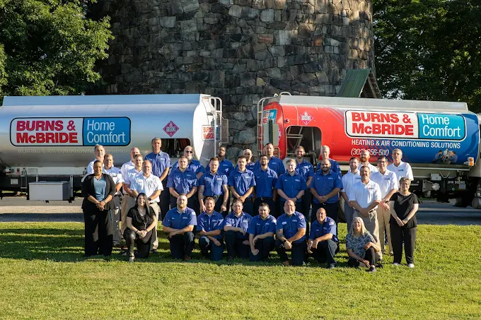 A group of people poses for a photo in front of two Burns & McBride Home Comfort fuel trucks, standing on grass with a stone structure in the background.